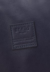 Étiquette en cuir marine avec le nom de la marque "anello" embossé et du texte. Présente une bordure cousue et une surface texturée.