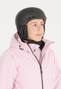 Black ski helmet with ventilation holes, paired with a light pink jacket featuring a high collar and zip closure. Smooth textures and modern design.