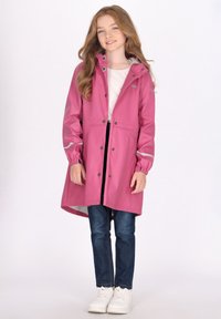 Rosa vattentät regnjacka med huva, tryckknappar och elastiska ärmar. Reflekterande detaljer och en slät textur. Bärs över en vit skjorta och jeans.