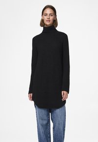 Schwarzer Rollkragenpullover mit lockerem Sitz, Rippenstruktur und langen Ärmeln. Wird über locker sitzenden blauen Jeans getragen.