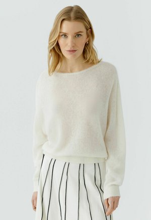 Frau mit blondem Haar, die einen weißen weichen Strickpullover und einen weißen Rock mit vertikalen schwarzen Streifen trägt, steht vor einem schlichten Hintergrund.