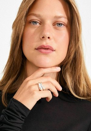 Hõberõngas lainelise, põimunud disainiga, taustal käsi lõual. Modell kannab musta pika varrukaga topi. Hele nahk ja juuksed.