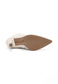 Zapato de tacón alto beige con punta afilada, textura de cuero suave y suela marrón sólida. Presenta un tacón delgado y detalles de costura sutiles.