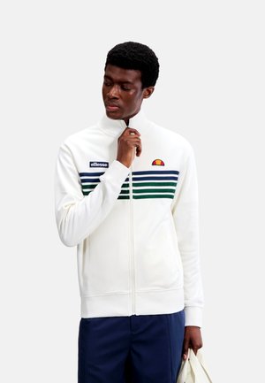 Hombre que lleva una chaqueta blanca con cremallera y rayas azules y verdes en el pecho, sujetando la cremallera con una mano y una bolsa blanca en la otra.