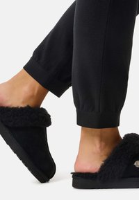 Stopy w czarnych, miękkich kapciach typu slip-on, zestawione z czarnymi spodniami z mankietami na jednolitym jasnym tle.
