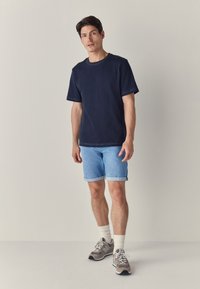 Maglietta di cotone blu navy con maniche corte, abbinata a pantaloncini di denim azzurro chiaro. Il modello indossa sneakers grigie e calzini bianchi a coste. Design semplice.