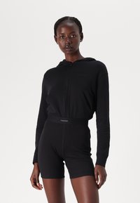 Svart zip-up hoodie med framsid ficka, i kombination med svart kort bodysuit. Båda har en figursydd design och Calvin Klein-logotyp på midjebandet.