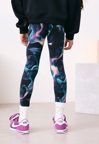 Színes leggings kék, lila és fekete örvénylő mintával; fehér zoknival és lila Nike tornacipővel párosítva; fekete hosszú ujjú pulóver.
