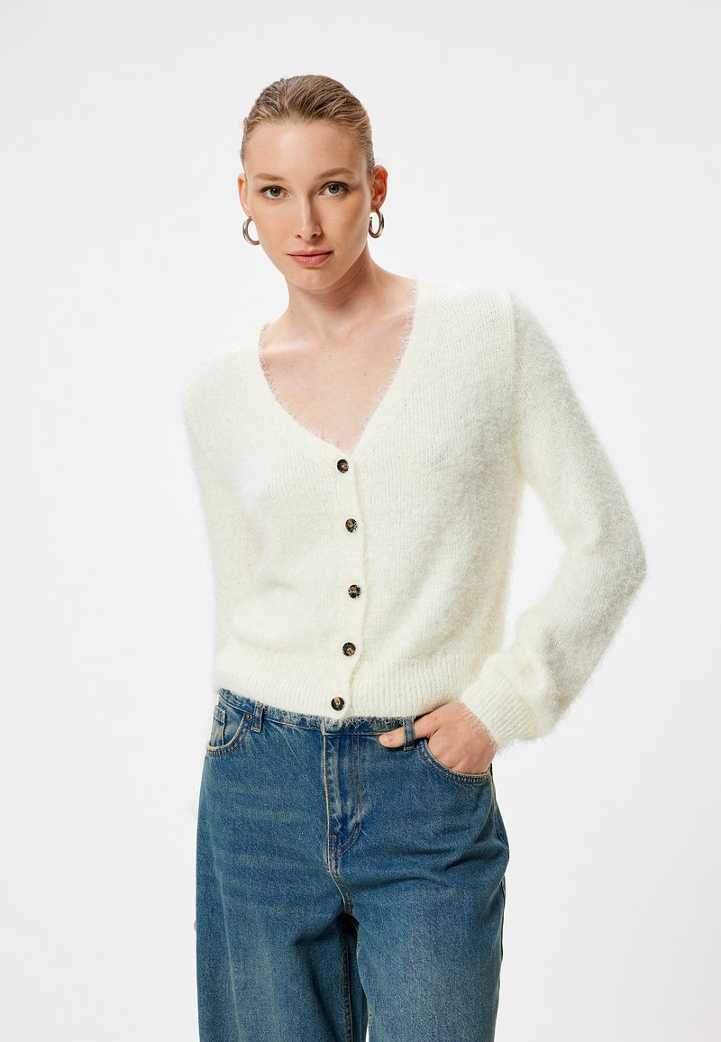 Cardigan bianco e soffice con scollo a V profondo, chiusura a bottoni e polsini a costine, abbinato a jeans in denim blu. La texture morbida è evidente.