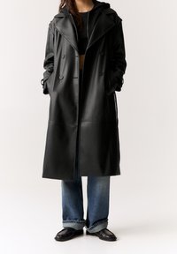 Czarny płaszcz typu trench z faux skóry, z podwójnym zapięciem, szerokimi klapami i regulowanymi rękawami. Nosi się go na czarnym crop topie i niebieskich dżinsach.