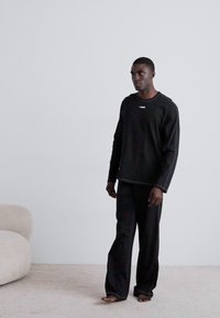Black long-sleeve top with contrasting neckline, paired with loose black pants. Soft fabric and simple design, standing on a light carpet.