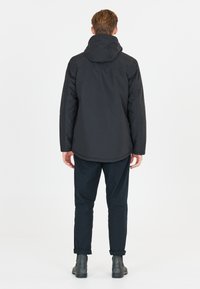 Svart vattentät jacka med huva, med en slät textur och en något längre baksida. Bärs med svarta byxor och stövlar.