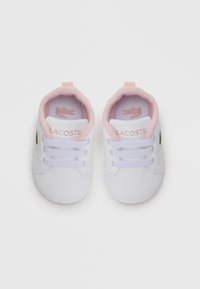 Bottines blanches pour bébé avec une surface lisse, semelle en caoutchouc, intérieur en maille rose et trois boucles en lacet blanches. Présente un petit logo vert sur le côté.
