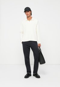 Cremfarbenes Langarm-Polohemd mit Kragen, kombiniert mit dunklen Hosen und schwarzen Schuhen. Das Modell trägt eine schwarze Tasche und eine schwarze Mütze.