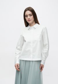 Nuori nainen, jolla on pitkät ruskeat hiukset, valkoinen napillinen paita ja vaaleansiniset laskostetut hame vasten yksinkertaista taustaa.