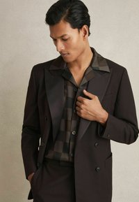Chaqueta de traje oscura de doble botón sobre una camisa de seda estampada. La camisa tiene un diseño de cuadrícula en negro y marrón, con botones y un cuello ancho.