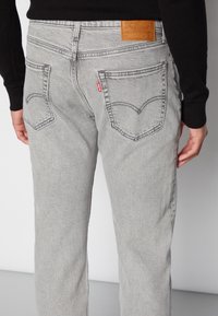 Harmaat denimit-farkut kapealla istuvuudella, joissa on kaksi takataskua ja nahkainen merkintä vyötäröllä. Saumat korostavat suunnittelua kontrastivärillään.