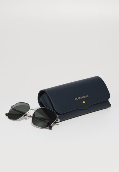 Occhiali da sole con lenti scure e montatura in metallo, forma angolata, affiancati da un astuccio in pelle blu navy con dettagli dorati e chiusura a scatto.