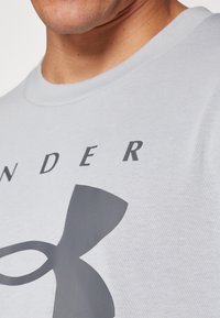 Lys grå t-skjorte med et stort mørk grått logodesign og fet tekst. Laget av myk stoff med rund hals og korte ermer.
