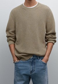 Mann trägt einen beigen Strickpullover über einem weißen Hemd mit Händen in den Taschen einer blauen Jeans, vor einem einfarbigen Hintergrund.