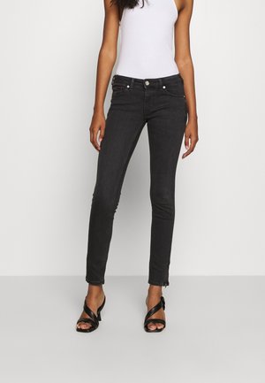 Frau trägt enge schwarze Jeans, weißes ärmelloses Oberteil und schwarze Sandalen mit Absatz, steht vor einem schlichten hellen Hintergrund.