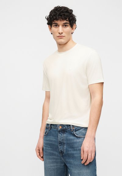 Νέος άνδρας με σγουρά μαλλιά, που φοράει απλό λευκό κοντομάνικο μπλουζάκι και μπλε τζιν, στέκεται μπροστά σε λευκό φόντο.