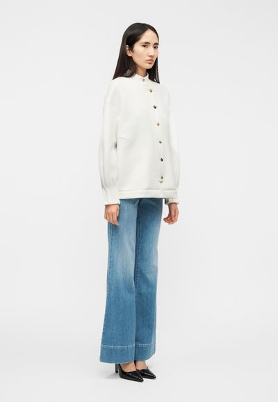 Λευκό φαρδύ φούτερ με ψηλό γιακά και χρυσά κουμπιά, σε συνδυασμό με ανοιχτό μπλε τζιν παντελόνι με φαρδύ πόδι και μαύρες μυτερές γόβες.
