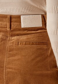 Pantalones de pana marrones con una textura suave, un bolsillo trasero y una etiqueta de tela blanca en la cintura. Detalles de costura con estrías sutiles.