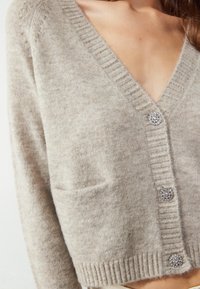 Cárdigan beige de punto con cuello en V, detalles acanalados, dos bolsillos y botones engastados. Textura suave y diseño casual.