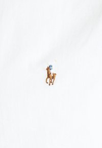 Bílá látka s malým hnědým vyšitým detailem koně a jezdce. Jezdec má modré tričko a drží žlutou hůlku.