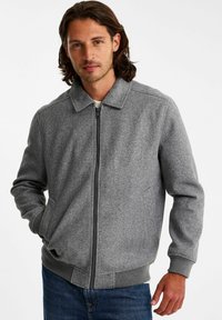 Grijze wolmix jas met een ritssluiting aan de voorkant, kraag, zijzakken en geribbelde manchetten. Draagt over een wit shirt met spijkerbroek.