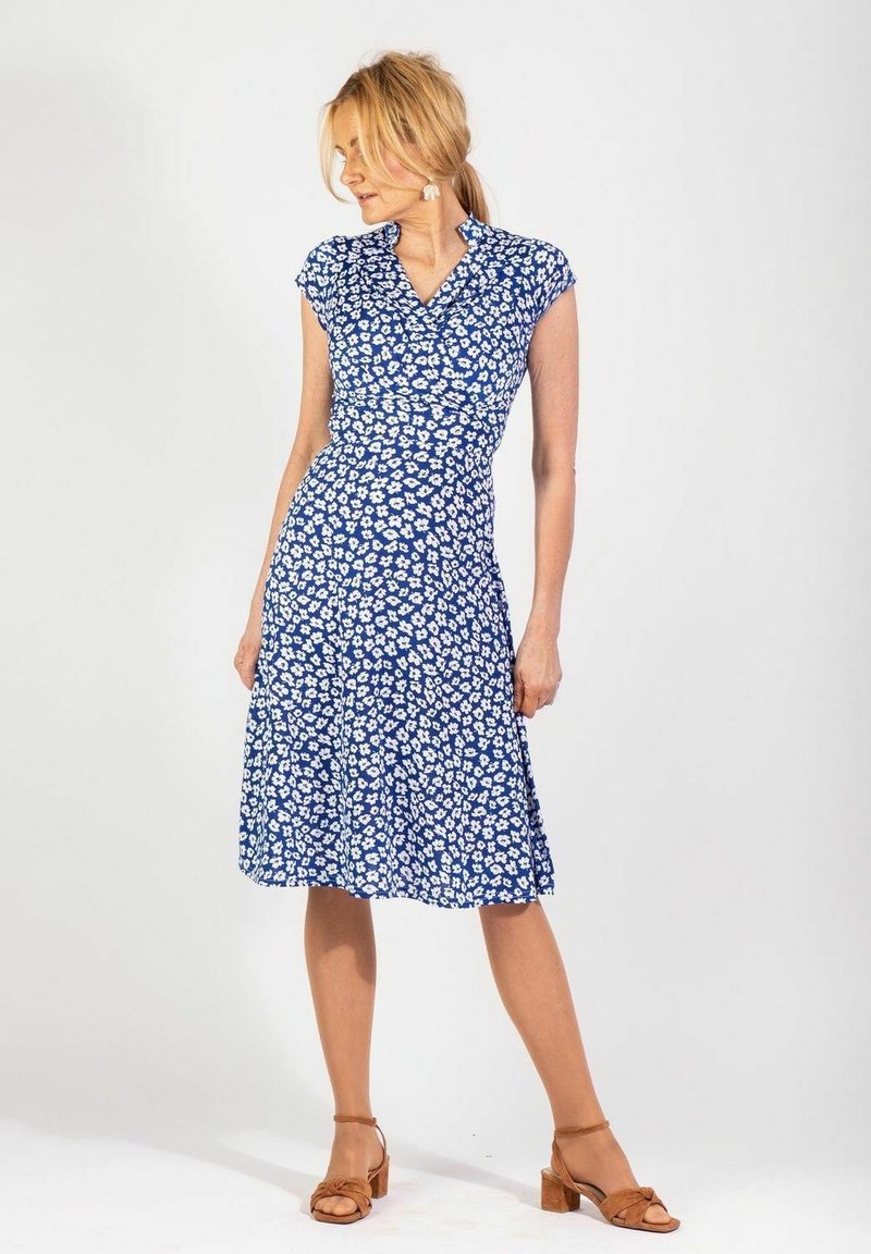 Sininen kukallinen mekko, jossa on lyhyet hihansuut, istuva yläosa ja levenevä helma. Kuosissa on pieniä valkoisia kukkia. Päällä ruskeat sandaalit.