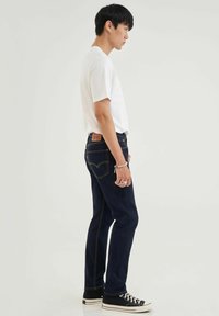Jeans de denim azul oscuro con costuras naranjas en contraste, corte ajustado y parche de cuero en la parte posterior. Llevados con una camiseta blanca básica y zapatillas negras.