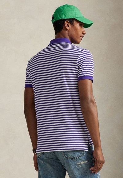 Άνδρας με πράσινο καπέλο, φορώντας ριγέ πόλο πουκάμισο μοβ και άσπρο και μπλε τζιν, στέκεται πλάγια σε απλό φόντο.