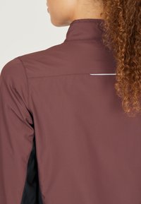 Veste de sport bordeaux avec un col haut, une texture lisse, et un accent réfléchissant dans le dos, avec des panneaux latéraux noirs.