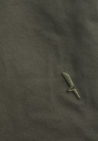 Dark green fabric with a small, embroidered olive green lightning bolt symbol near the bottom right corner.