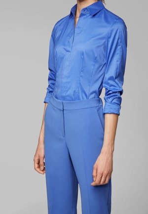 Personne portant une chemise en satin bleu ajustée à manches longues rentrée dans un pantalon bleu taille haute sur mesure, avec les manches retroussées.