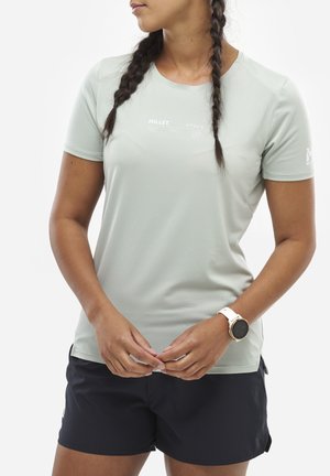 Femme aux cheveux tressés portant un t-shirt Millet gris clair, un short noir et une montre connectée blanche, debout devant un fond blanc.