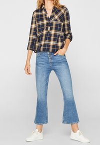 Námořnická modrá a žlutá kostkovaná košile na tlačítka s tříčtvrtečními rukávy, kombinovaná se světle modrými rozšířenými džíny a bílými teniskami.