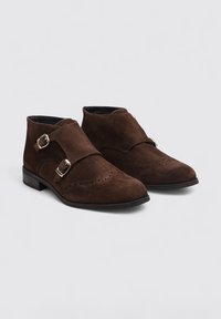 Botas de tobillo de ante marrón con un diseño de doble hebilla, punta afilada y detalles perforados a lo largo del borde superior.
