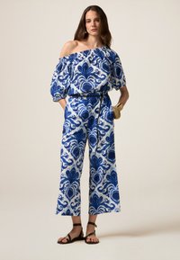 Blauw-wit gestreepte jumpsuit met een off-shoulder ontwerp, wijde mouwen en korte benen, cinched in de taille met een zwarte riem.