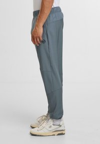 Grå idrottsbyxor med en slät textur, elastisk midja, sidofickor och en subtil logotypdetalj på låret. Bärs med vita sneakers.