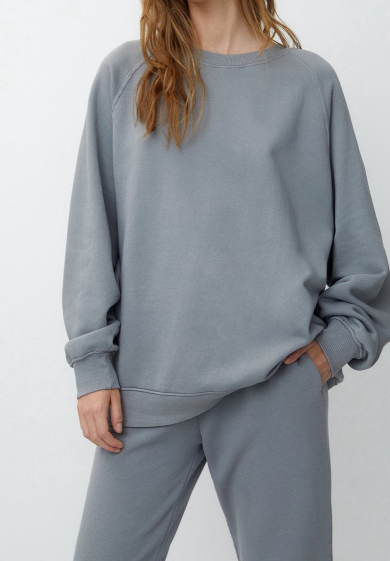 Femme portant un sweat-shirt gris clair ample et un pantalon de survêtement assorti, une main dans la poche et un fond neutre.