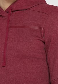 Sweat à capuche rouge avec tissu texturé, cordon de serrage réglable et détail de poche poitrine brillante. Finition lisse, coupe ajustée avec un look moderne.