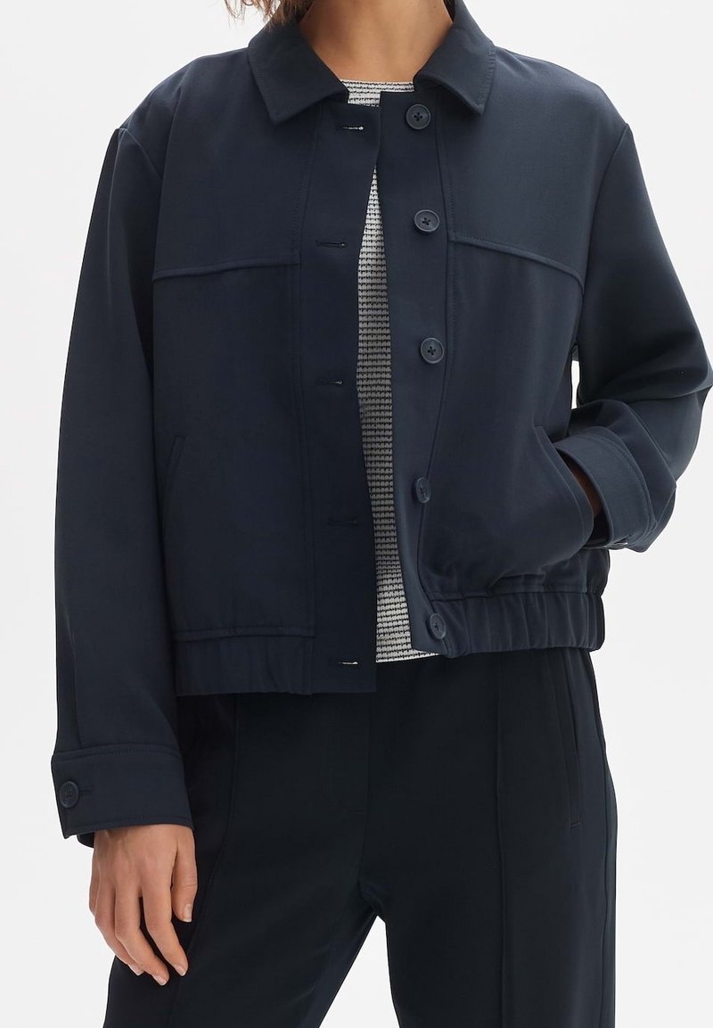 Laivastonsininen takki, joka on valmistettu sileästä kankaasta. Siinä on nappi-edessä suljenta, kaulus ja joustava vyötärö. Muotoiltu malli, jossa on sivutaskut.