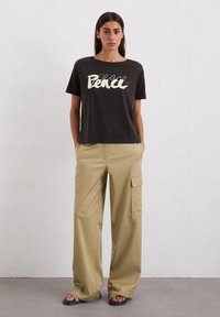 Schwarzes Baumwoll-T-Shirt mit weißem "Frieden"-Grafik, kombiniert mit hellbeigen, weit geschnittenen Cargo-Hosen mit Seitentaschen und flachen schwarzen Sandalen.