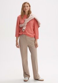 Korallenwollpullover mit Rundhalsausschnitt, kombiniert mit beigen, weit geschnittenen Hosen. Hellrosa strukturierten Strickwrap, der über dem Pullover drapiert ist. Weiße Sneakers.