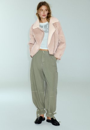 Femme portant une veste en peau retournée rose, un top court bleu clair, un pantalon ample vert olive noué aux chevilles et des chaussures noires sans lacets.
