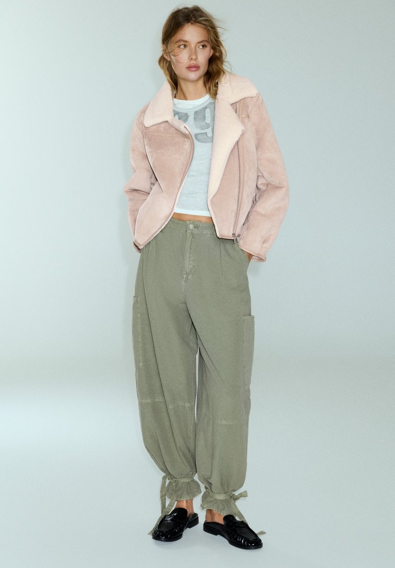 Femme portant une veste en peau retournée rose, un top court bleu clair, un pantalon ample vert olive noué aux chevilles et des chaussures noires sans lacets.