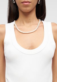 Mujer con top blanco sin mangas, collar de perlas y pendientes de aro plateados, con el cabello oscuro y liso visible sobre los hombros.